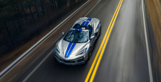 Overhead front view of 2024 Chevrolet Corvette E-Ray 3LZ convertible in Silver Flare with Electric Blue stripe package driving on a road between trees. Pre-production model shown. Actual production model may vary. Model year 2024 Corvette E-Ray available 2023.