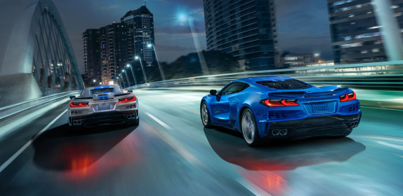 Rear view of 2024 Chevrolet Corvette E-Ray 3LZ coupe in Riptide Blue and convertible in Silver Flare with Electric Blue stripe package driving across a city bridge at night. Pre-production model shown. Actual production model may vary. Model year 2024 Corvette E-Ray available 2023.