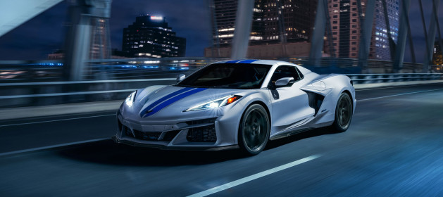 Front 3/4 view of 2024 Chevrolet Corvette E-Ray 3LZ convertible in Silver Flare with Electric Blue stripe package driving across a city bridge at night. Pre-production model shown. Actual production model may vary. Model year 2024 Corvette E-Ray available 2023.