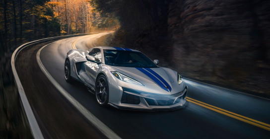 Passenger side front 3/4 view of 2024 Chevrolet Corvette E-Ray 3LZ convertible in Silver Flare with Electric Blue stripe package driving on a curved mountain road. Pre-production model shown. Actual production model may vary. Model year 2024 Corvette E-Ray available 2023.